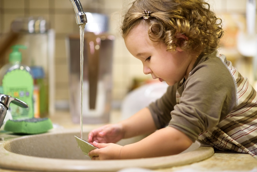 ute two-year-old girl is washing dishes in the kitchen. Baby helps the parents. Nice little child learns the household. Toddler washes the plate in the sink. Adorable baby does house cleaning.