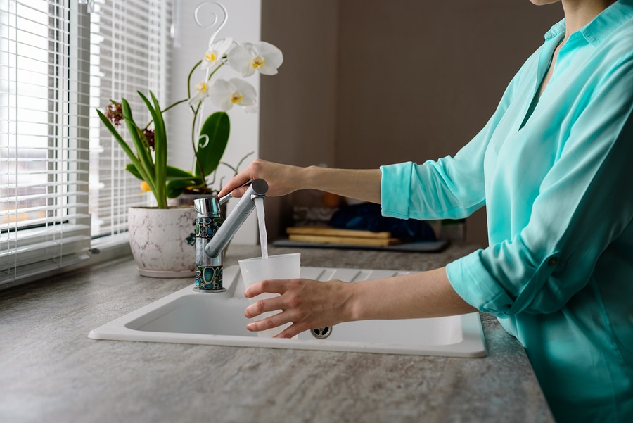 close-up of a woman collects water in a plastic glass from the tap in the kitchen sink in front of the window.